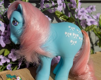 Peut inclure: Un poney jouet bleu clair vintage avec des cheveux et une queue roses. Le poney a des accents de nœuds blancs sur le côté. Le poney est posé sur une surface en bois avec des fleurs violettes en arrière-plan. Le poney a un logo sur son pied.
