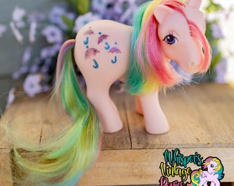 May include: A light pink plastic toy pony with a long, flowing mane and tail featuring streaks of yellow, green, and pink. The pony has purple eyes and a glittery blue and pink umbrella pattern on its flank. A logo reads Whisper's Vintage Ponies.