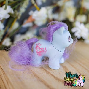May include: A light blue toy pony with purple hair and tail. A pink cupcake graphic is on the pony's side. The pony has pink eyes and is on a wooden surface. The logo "Whispers Vintage Ponies" is in the bottom right corner.