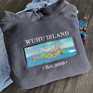 Puede incluir: Una sudadera con capucha gris oscuro con un dise&ntilde;o bordado en blanco que dice "WUHU ISLAND" y una peque&ntilde;a imagen bordada de una isla con un edificio en ella. El texto "Est. 2009" tambi&eacute;n est&aacute; bordado en la sudadera.