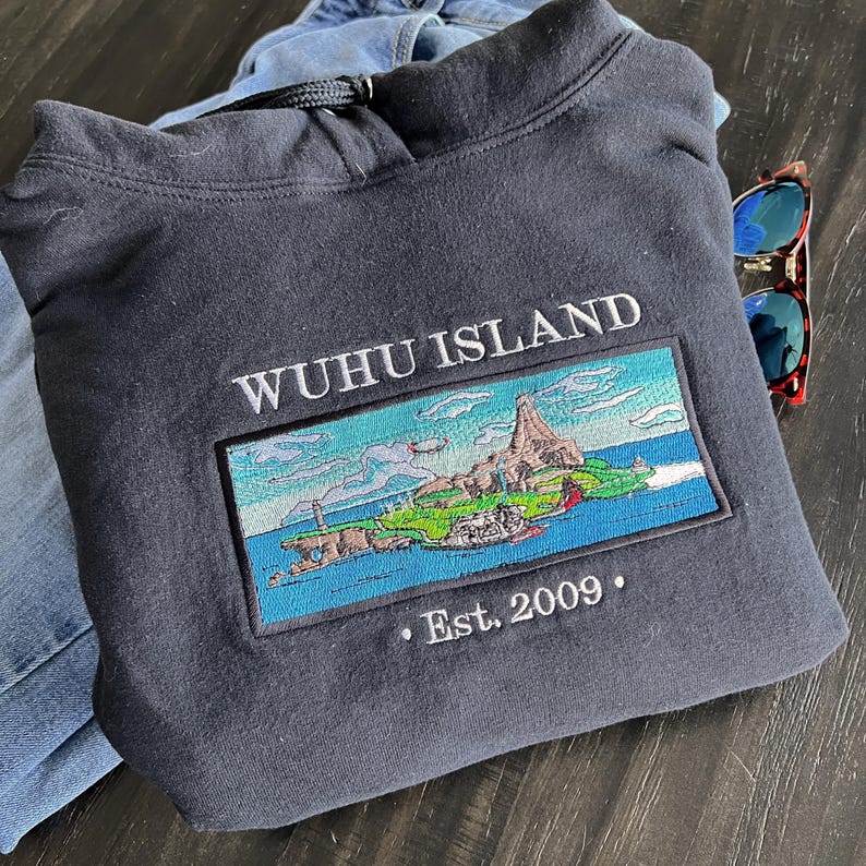 Puede incluir: Una sudadera con capucha gris oscuro con un dise&ntilde;o bordado en blanco que dice "WUHU ISLAND" y una peque&ntilde;a imagen bordada de una isla con un edificio en ella. El texto "Est. 2009" tambi&eacute;n est&aacute; bordado en la sudadera.