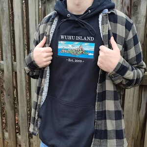 Puede incluir: Una sudadera con capucha negra con un gr&aacute;fico bordado blanco de una isla con el texto "WUHUI ISLAND" y "Est. 2009". La sudadera se lleva sobre una camisa de franela a cuadros.