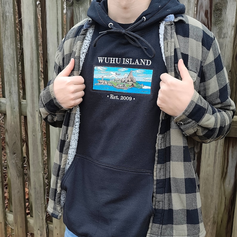 Puede incluir: Una sudadera con capucha negra con un gr&aacute;fico bordado blanco de una isla con el texto "WUHUI ISLAND" y "Est. 2009". La sudadera se lleva sobre una camisa de franela a cuadros.