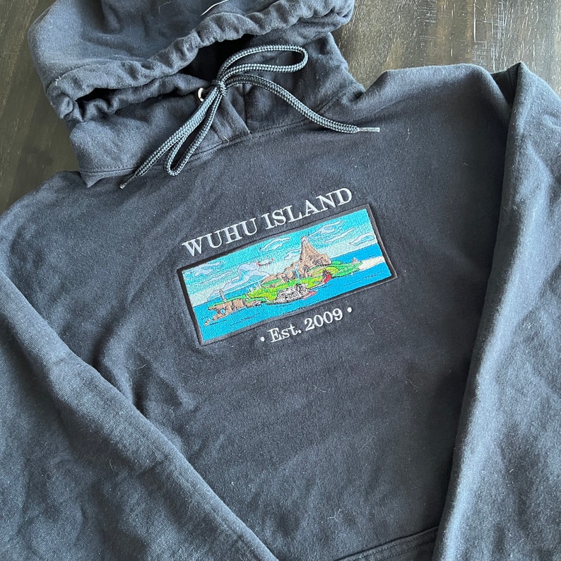 Puede incluir: Sudadera con capucha negra con una imagen bordada en blanco de una isla con el texto "WUHU ISLAND . Est. 2009."