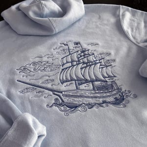 Könnte beinhalten: Hellblauer Kapuzenpullover mit einem gestickten Design eines Segelschiffs auf dem Meer. Das Schiff hat mehrere Segel und ist von stilisierten Wolken und Wellen umgeben. Der Hoodie hat eine Kapuze und eine Vordertasche.