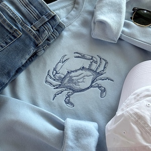 Może przedstawiać: Jasno błękitna bluza z haftowanym wzorem niebieskiego kraba. Bluza jest złożona i leży na powierzchni z niebieskimi dżinsami i białą czapką bejsbolową.