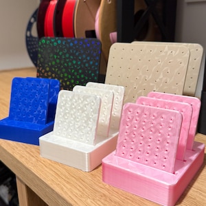 May include: A collection of 3D printed earring display stands in blue, white, and pink, each featuring a rectangular base and a vertical panel with a geometric faceted surface pattern and multiple small holes for organizing jewelry pieces.