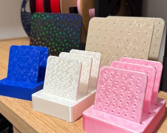 May include: A collection of 3D printed earring display stands in blue, white, and pink, each featuring a rectangular base and a vertical panel with a geometric faceted surface pattern and multiple small holes for organizing jewelry pieces.