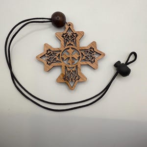May include: A wooden cross pendant with intricate cut-out designs and a central cross motif. The pendant is attached to a black cord with a wooden bead and a black adjustable toggle. The cross is approximately 3 inches in diameter.