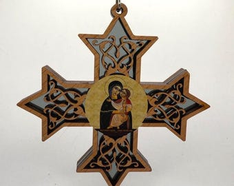 May include: A wooden cross ornament with a detailed, intricate design. The cross features a circular image of the Virgin Mary and baby Jesus in the center. The cross is painted in shades of brown, black, and gold.