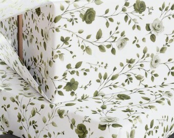 Può includere: Due scatole regalo avvolte in carta bianca con un motivo floreale verde. Le scatole sono impilate l'una sull'altra. Sullo sfondo sono visibili un paio di forbici e un rotolo di carta da regalo. Un nastro grigio è in primo piano.