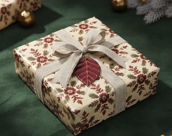 May include: A wrapped gift box with floral patterned paper in cream, burgundy, and green. A gray ribbon and bow secure the package, with a burgundy leaf-shaped charm. The gift sits on a dark green surface, with gold ornaments and greenery in the background.