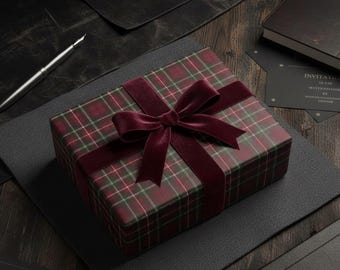 May include: A rectangular gift box wrapped in dark red and green plaid paper, tied with a wide, deep burgundy velvet ribbon bow. The box is positioned on a dark surface alongside a silver fountain pen and black invitation cards.