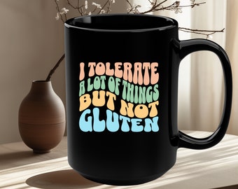 Puede incluir: Taza de café negra con un diseño blanco que dice "Tolero muchas cosas pero no el gluten" en un arcoíris de colores.