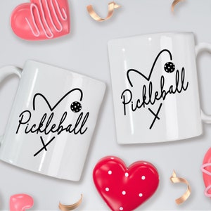 May include: Two white coffee mugs with black hearts and the text "Pickleball" on them. The mugs are surrounded by pink and red hearts.