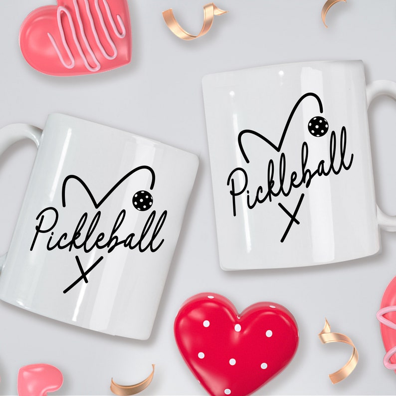 May include: Two white coffee mugs with black hearts and the text "Pickleball" on them. The mugs are surrounded by pink and red hearts.