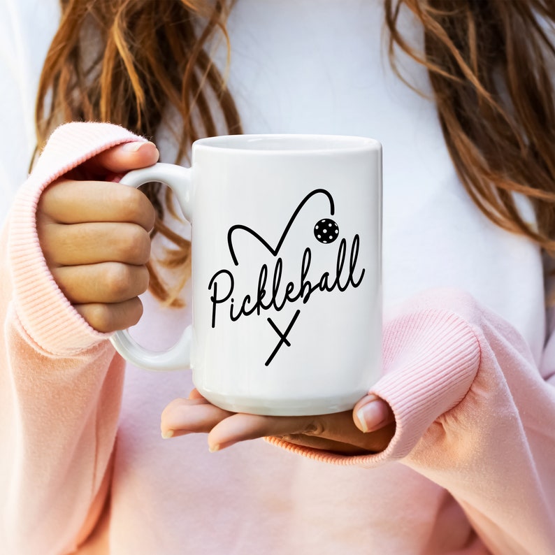 May include: White ceramic coffee mug with a black graphic of a heart with a pickleball paddle and ball inside and the text "Pickleball" written in cursive.