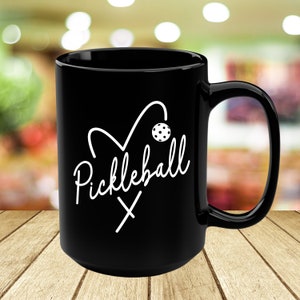 May include: Black coffee mug with a white design of a heart with a pickleball paddle and ball inside. The text "Pickleball" is written in a cursive font.