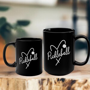 May include: Two black coffee mugs with a white design. The design features a heart with a pickleball paddle and ball inside. The text "Pickleball" is written in cursive below the heart.