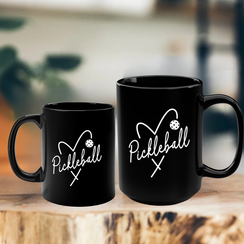 May include: Two black coffee mugs with a white design. The design features a heart with a pickleball paddle and ball inside. The text "Pickleball" is written in cursive below the heart.