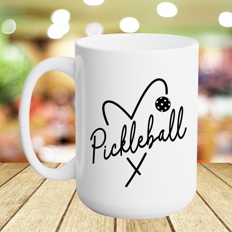 May include: White ceramic coffee mug with a black and white graphic of a heart with a pickleball paddle and ball. The text "Pickleball" is written in a cursive font.