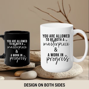 May include: Two white ceramic mugs with black text. The text on the mugs reads "You are allowed to be both a masterpiece & a work in progress". The mugs are on a wooden surface with a branch in the background.