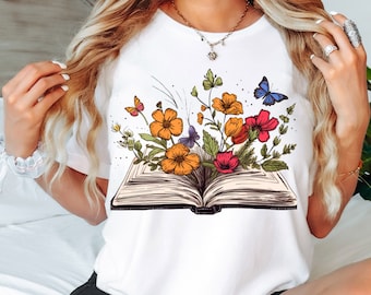 May include: White t-shirt featuring a graphic of an open book with colourful flowers and butterflies emerging from the pages.
