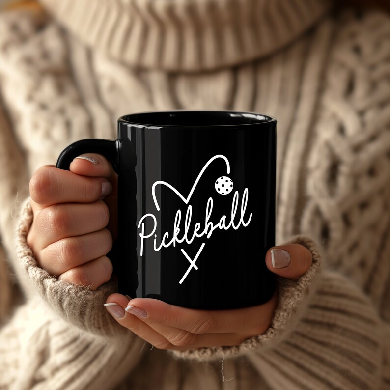 May include: A black coffee mug with a white heart design and the text "Pickleball" in a cursive font. The heart design includes a pickleball paddle and ball.