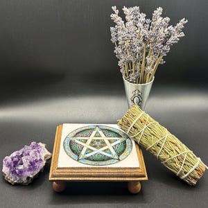 May include: A collection of spiritual items on a black surface. Includes a wooden stand with a pentagram design, a bundle of sage, a cluster of amethyst crystals, and a small metal vase with lavender.