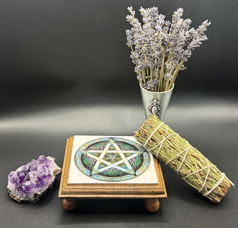 May include: A collection of spiritual items on a black surface. Includes a wooden stand with a pentagram design, a bundle of sage, a cluster of amethyst crystals, and a small metal vase with lavender.