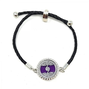 May include: A black leather bracelet with a silver charm featuring a tree of life design with a purple center and clear rhinestones around the edge.