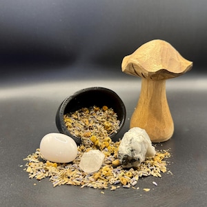 May include: A wooden mushroom carving stands next to a small black bowl filled with dried herbs and flowers. Two clear quartz crystals and a pink stone are scattered around the bowl. The background is black.