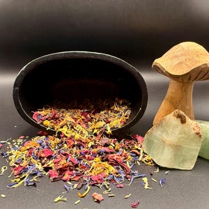 May include: A black ceramic bowl tipped over, spilling a colorful mix of dried flower petals. A carved wooden mushroom and two green stones are in the background. The petals are red, yellow, blue, and orange.
