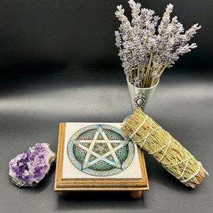 May include: A decorative wooden stand with a tile featuring a pentagram design, a bundle of dried herbs, a cluster of purple amethyst crystals, and a metal vase with lavender sprigs. The items are arranged on a dark surface.