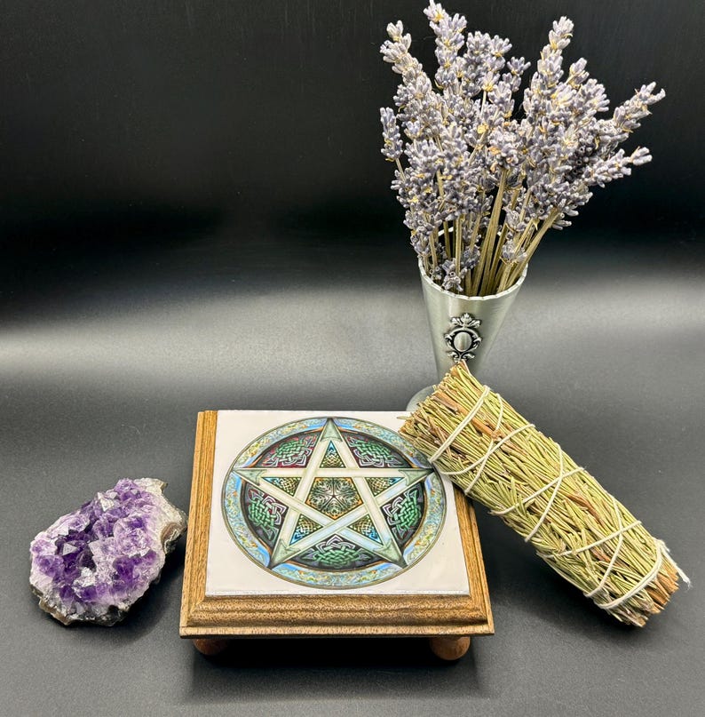 May include: A decorative wooden stand with a tile featuring a pentagram design, a bundle of dried herbs, a cluster of purple amethyst crystals, and a metal vase with lavender sprigs. The items are arranged on a dark surface.