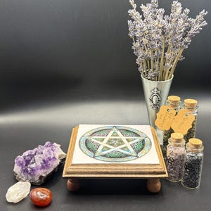 May include: A wooden stand with a tile top featuring a colorful pentagram design. The stand is surrounded by amethyst crystals, small glass jars with cork stoppers, and a metal vase with lavender. A red and clear stone are in front.
