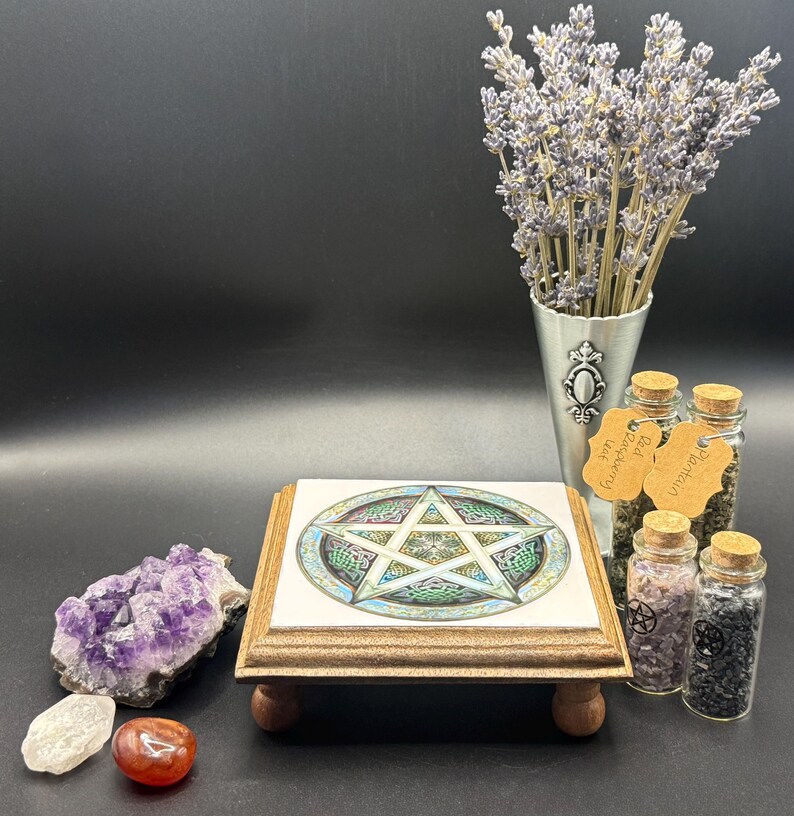 May include: A wooden stand with a tile top featuring a colorful pentagram design. The stand is surrounded by amethyst crystals, small glass jars with cork stoppers, and a metal vase with lavender. A red and clear stone are in front.