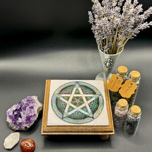 May include: A wooden altar tile with a pentagram design, featuring green, blue, and gold accents. The tile is surrounded by crystals, herbs in glass jars, and a bouquet of lavender. The overall theme is spiritual and mystical.