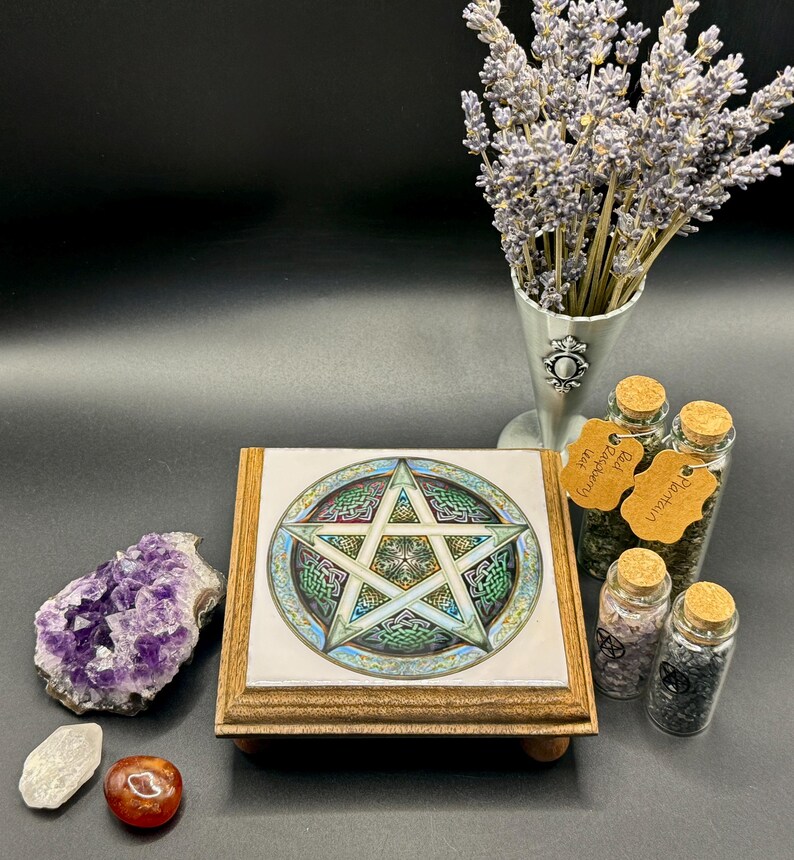 May include: A wooden altar tile with a pentagram design, featuring green, blue, and gold accents. The tile is surrounded by crystals, herbs in glass jars, and a bouquet of lavender. The overall theme is spiritual and mystical.