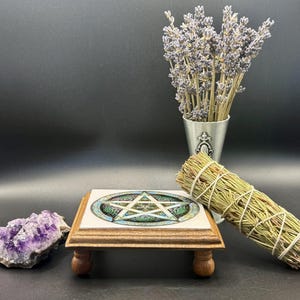 May include: A wooden altar tile with a pentagram design, a bundle of sage, a purple amethyst crystal, and a small metal vase with lavender. The altar tile has a white tile top with a brown wooden frame and four small wooden feet.