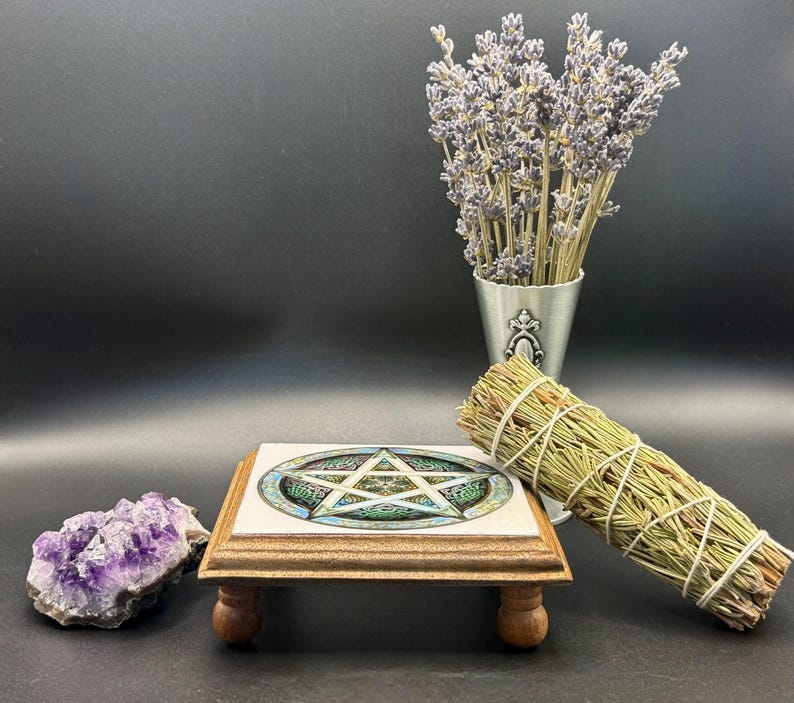 May include: A wooden altar tile with a pentagram design, a bundle of sage, a purple amethyst crystal, and a small metal vase with lavender. The altar tile has a white tile top with a brown wooden frame and four small wooden feet.