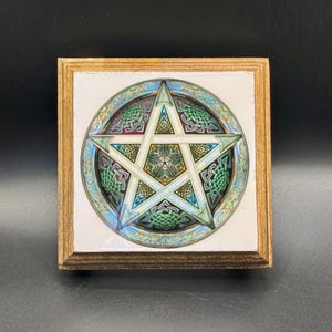 May include: A square wooden plaque featuring a pentagram design. The pentagram is set within a circular border with Celtic knot patterns in green, blue, and white. The wooden frame has a light brown finish.