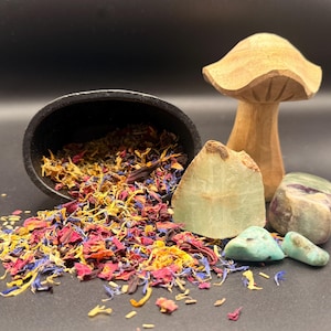 May include: A still-life arrangement featuring a wooden mushroom, various gemstones, and a black bowl overflowing with colorful dried flower petals. The gemstones include green and turquoise stones. The background is black.