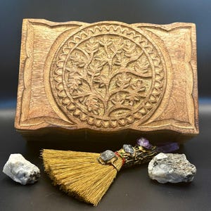 May include: A rectangular wooden box with a carved tree of life design on the lid. A small broom, crystals, and stones are arranged in front of the box. The box and broom are light brown, and the crystals are gray and purple.