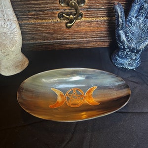 May include: A light brown, oval-shaped dish made of horn with a gold pentacle and triple moon symbol etched into the surface.