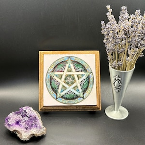 May include: A framed artwork featuring a detailed pentagram design with Celtic knot patterns in green, blue, and white. A purple amethyst geode and a silver vase with lavender sprigs are also present.
