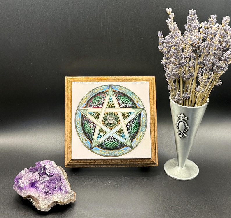 May include: A framed artwork featuring a detailed pentagram design with Celtic knot patterns in green, blue, and white. A purple amethyst geode and a silver vase with lavender sprigs are also present.