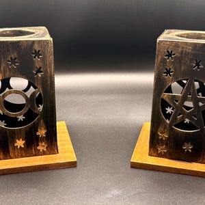 May include: Two rectangular candle holders with a distressed black and gold finish, each mounted on a square wooden base. One holder features a triple moon cutout, while the other displays a pentagram cutout, both accented with small star-shaped perforations.