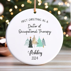 May include: A round white ceramic Christmas ornament hanging by a gold string. The text reads First Christmas as a Doctor of Occupational Therapy, Ashley, and 2024. Below the text are five stylized Christmas tree illustrations in green, gold, and black.