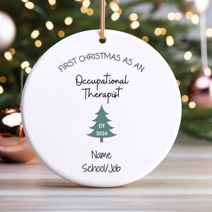May include: A round white ceramic ornament hanging by a gold string. The text reads FIRST CHRISTMAS AS AN Occupational Therapist, with a green Christmas tree graphic featuring the text OT 2024, followed by the placeholder text Name School/Job.